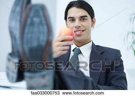 Young professional man sitting with feet up, holding apple and smiling at camera View Large Photo Image Stock Image - Young professional man sitting with feet up, holding apple and smiling at camera. Fotosearch