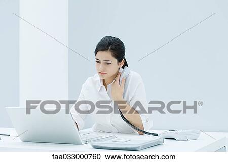 Stock Image - Young professional woman sitting at desk, using laptop, holding landline phone to her chest. Fotosearch