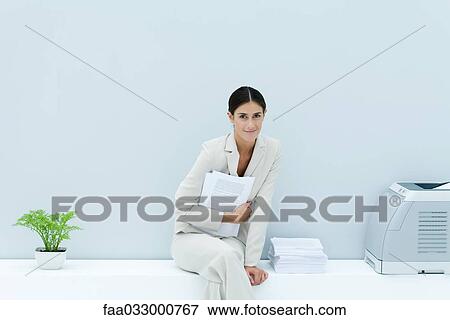 Stock Photo - Young professional woman sitting on ledge with legs crossed, holding documents and smiling at camera. Fotosearch