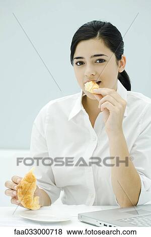 Stock Photo - Young woman eating croissant, sitting at desk, looking at camera. Fotosearch
