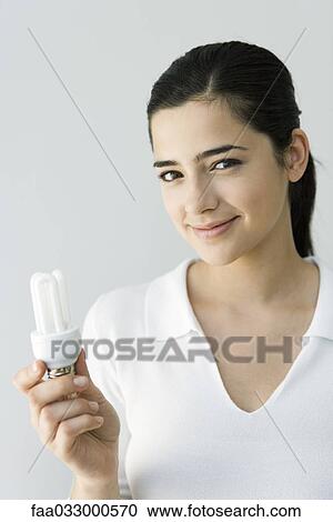 Stock Image - Young woman holding energy efficient light bulb, smiling at camera. Fotosearch