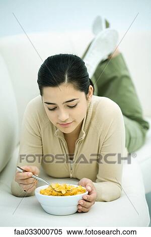 Young woman lying on stomach on couch, holding cereal bowl, looking down View Large Photo Image Stock Photography - Young woman lying on stomach on couch, holding cereal bowl, looking down. Fotosearch