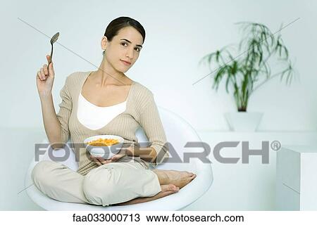Stock Image - Young woman sitting in chair, holding cereal bowl and spoon, smiling at camera. Fotosearch