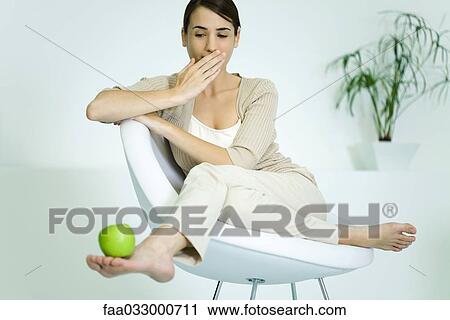 Stock Image - Young woman slouching in chair, balancing apple on foot, covering mouth. Fotosearch