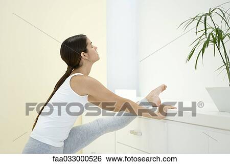 Stock Photograph - Young woman stretching leg against chest of drawers. Fotosearch