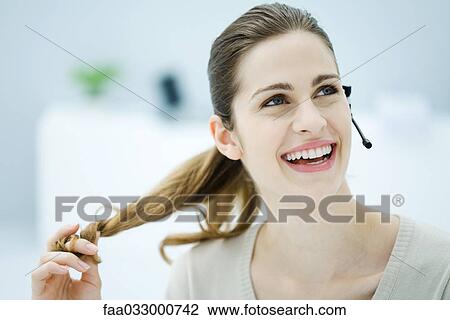 Stock Image - Young woman twirling her hair, wearing headset and looking away, smiling. Fotosearch