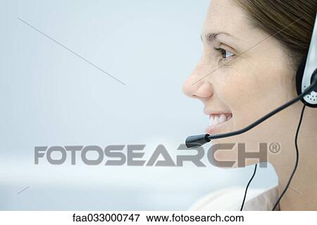Stock Photo - Young woman wearing headset, looking away and smiling, side view. Fotosearch