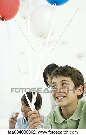 Stock Image - Boy holding balloons, smiling, family in background. Fotosearch