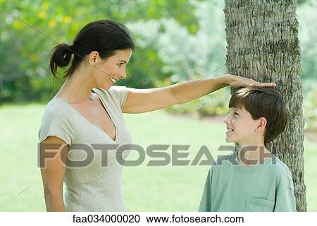 Stock Image - Boy standing against tree trunk, his mother placing her hand on his head, both smiling looking at each other. Fotosearch
