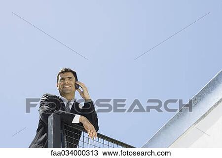 Stock Image - Businessman leaning against railing, using cell phone, low angle view. Fotosearch