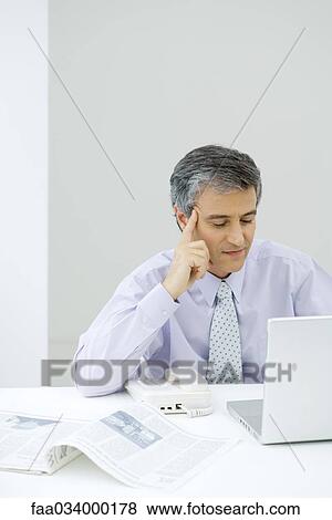 Businessman sitting at desk, looking at laptop computer, holding head View Large Photo Image Stock Photo - Businessman sitting at desk, looking at laptop computer, holding head. Fotosearch