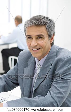 Stock Image - Businessman sitting in office, smiling at camera, portrait. Fotosearch