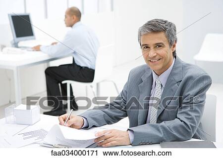 Stock Image - Businessman working at desk, smiling at camera, colleague in background. Fotosearch