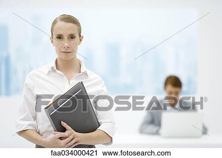 Stock Image - Businesswoman holding folder, looking at camera, male colleague in background. Fotosearch