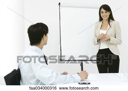 Stock Photograph - Businesswoman making presentation, smiling at camera, male colleague taking notes. Fotosearch