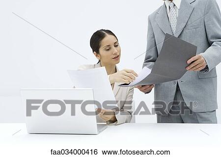 Stock Photograph - Businesswoman sitting at desk, discussing document with male colleague. Fotosearch