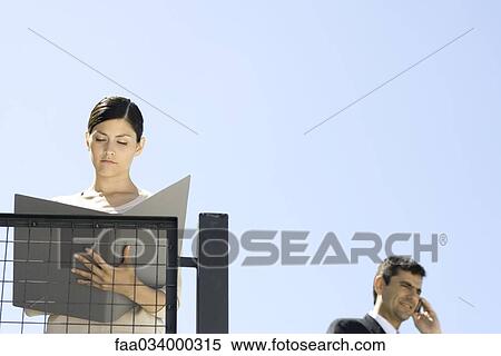 Stock Photography - Businesswoman standing outdoors, looking at document, colleague using cell phone in background. Fotosearch