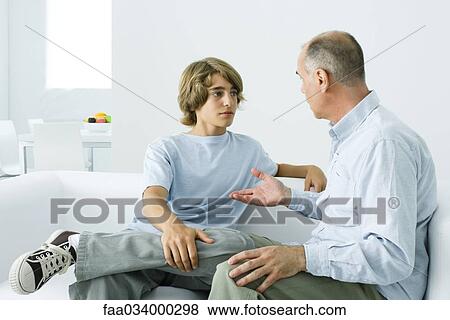 Stock Photo - Father and teen son sitting together on sofa, having discussion. Fotosearch