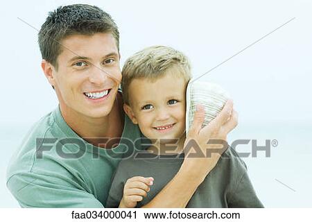 Stock Image - Father holding seashell to son's ear, both smiling, portrait. Fotosearch