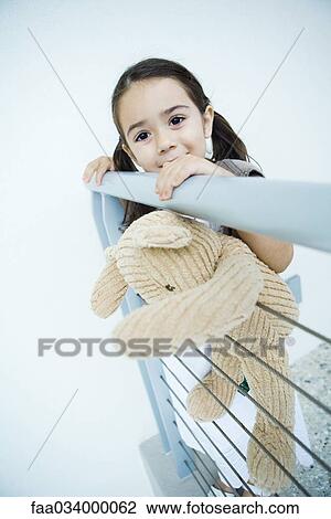Stock Image - Little girl standing at rail holding teddy bear, smiling at camera. Fotosearch
