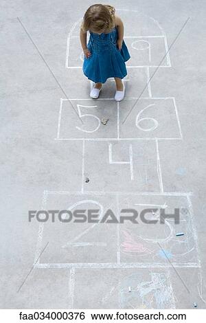 Stock Photograph - Little girl standing on hopscotch grid, looking down at rock, high angle view. Fotosearch