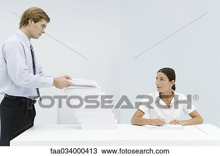 Stock Image - Man adding to large stack of documents on professional woman's desk. Fotosearch