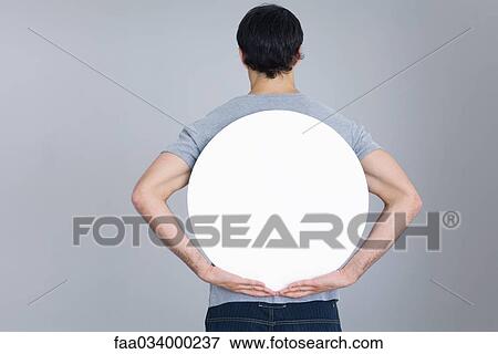 Stock Photo - Man holding blank circular sign behind back, rear view. Fotosearch