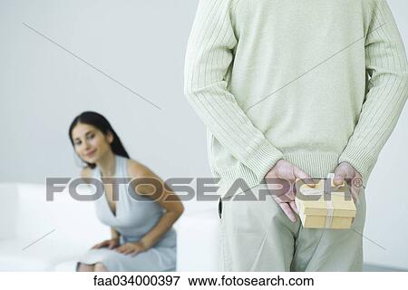 Stock Photo - Man holding gift behind his back, woman trying to peek in background, cropped view. Fotosearch