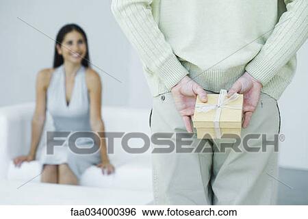 Stock Photograph - Man holding gift behind his back, woman smiling in background, cropped view. Fotosearch