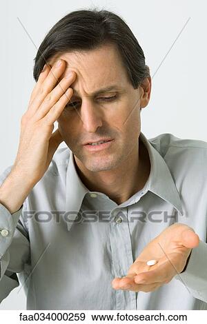 Stock Photo - Man holding head, looking down at pill in hand. Fotosearch