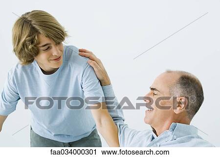 Stock Image - Man sitting with hand on teen son's shoulder, both smiling. Fotosearch