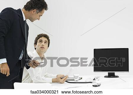 Stock Image - Professional man at desk holding phone, looking up at manager standing beside him. Fotosearch