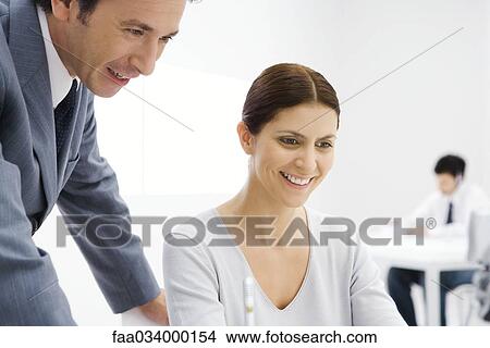 Picture - Professional man leaning over woman's shoulder, both looking down and smiling. Fotosearch