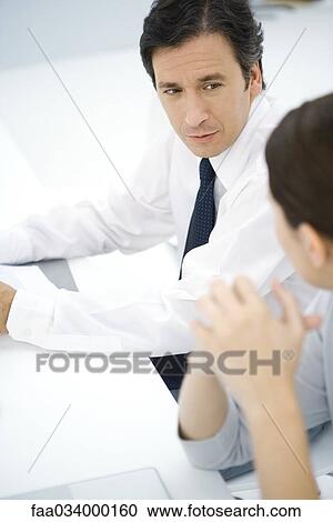 Stock Image - Professional man sitting at desk, talking with female colleague, high angle view. Fotosearch
