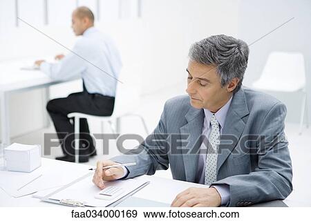 Stock Photo - Professional man sitting at desk, using calculator, colleague in background. Fotosearch