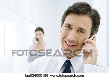 Professional man using cell phone, smiling at camera, woman on phone in background View Large Photo Image Stock Photo - Professional man using cell phone, smiling at camera, woman on phone in background. Fotosearch