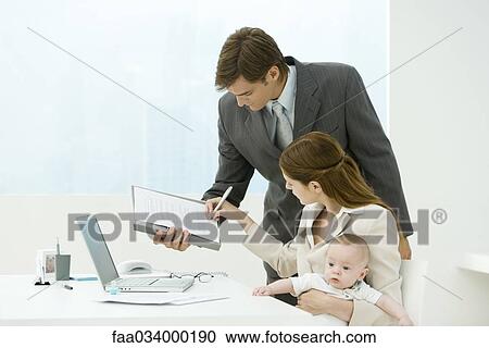 Stock Image - Professional woman signing document in office, holding baby on lap. Fotosearch