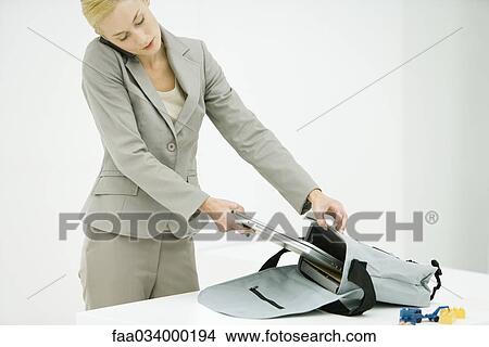 Picture - Professional woman using cell phone, putting laptop computer in briefcase, child's toys nearby. Fotosearch