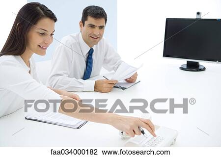 Stock Image - Professionals sitting at table, woman dialing landline phone. Fotosearch