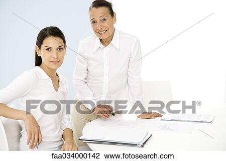 Stock Image - Senior woman and younger colleague discussing document, both smiling at camera. Fotosearch