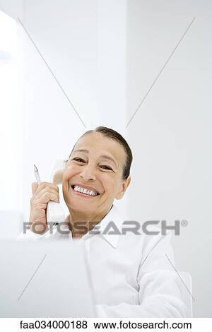 Stock Photo - Senior woman using landline phone and laptop computer, smiling at camera. Fotosearch