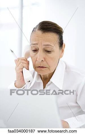 Senior woman using phone, frowning at laptop computer View Large Photo Image Stock Photo - Senior woman using phone, frowning at laptop computer. Fotosearch