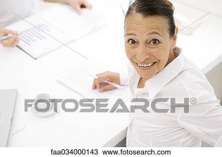 Senior woman working at table, smiling up at camera View Large Photo Image Stock Image - Senior woman working at table, smiling up at camera. Fotosearch