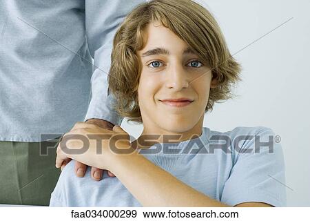 Stock Photo - Teen boy holding father's hand on shoulder, smiling at camera, cropped view. Fotosearch