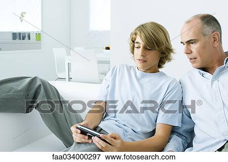 Stock Photo - Teen boy playing handheld video game, father watching over his shoulder. Fotosearch