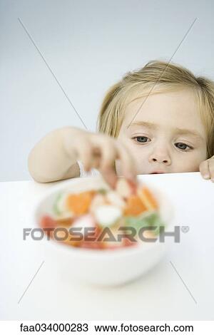 Stock Image - Toddler girl reaching for bowl of candy. Fotosearch