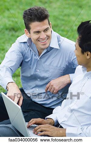 Two men sitting in park with laptop computer, looking at each other smiling View Large Photo Image Stock Photograph - Two men sitting in park with laptop computer, looking at each other smiling. Fotosearch