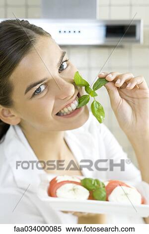 Stock Photography - Woman biting into sprig of basil, holding plate of Caprese salad. Fotosearch