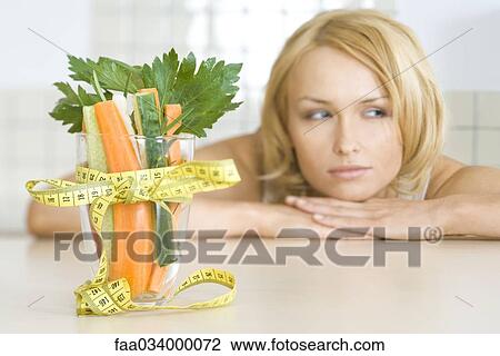 Stock Image - Woman contemplating glass filled with fresh vegetables wrapped with measuring tape tied in bow. Fotosearch