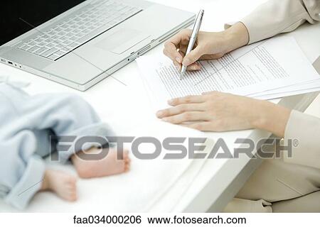 Stock Photograph - Woman editing document at desk, infant lying nearby, cropped view. Fotosearch
